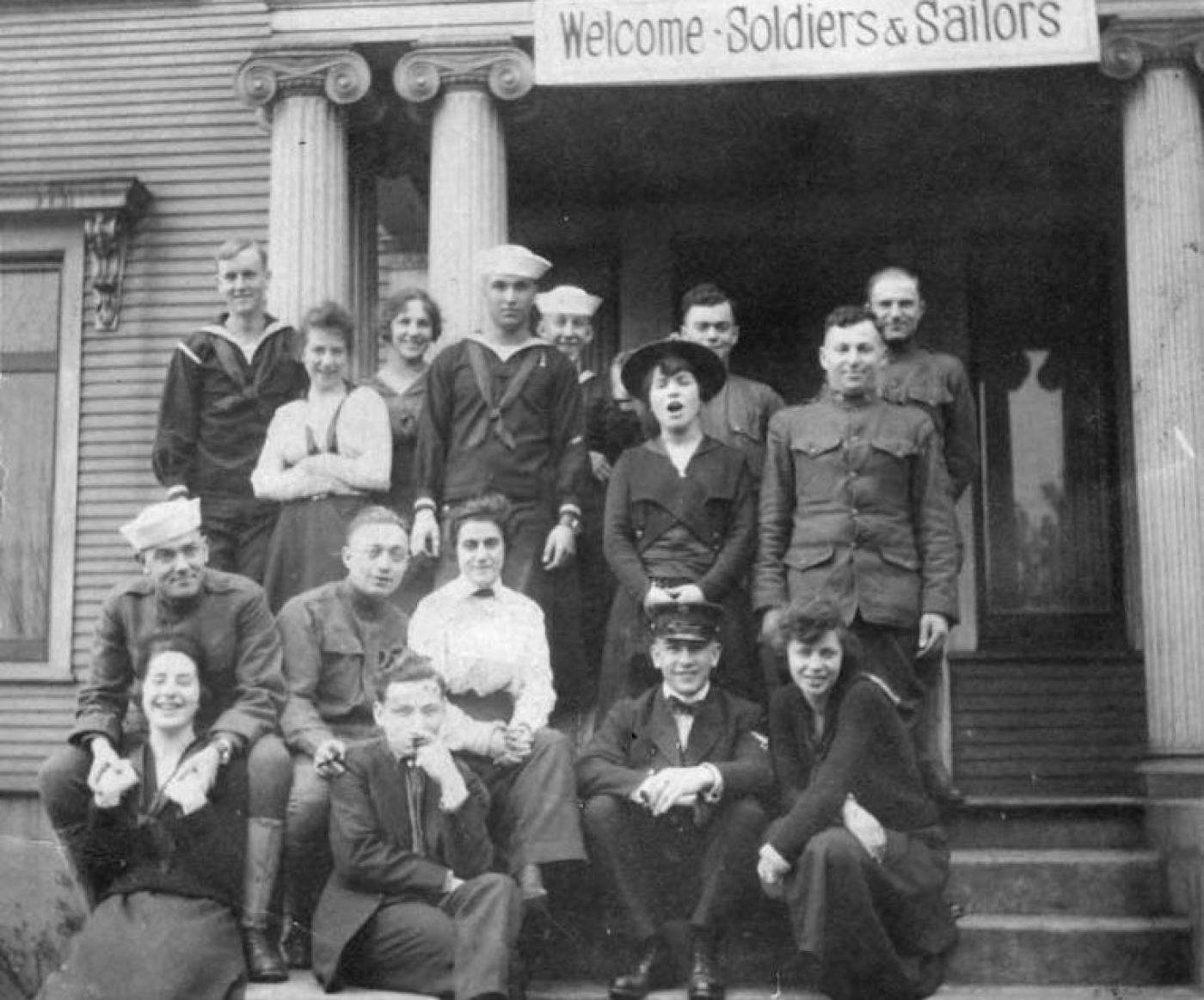 Group of women and men, some in military uniforms, on steps of house with sign above door reading "Welcome Soldiers & Sailors", 1921