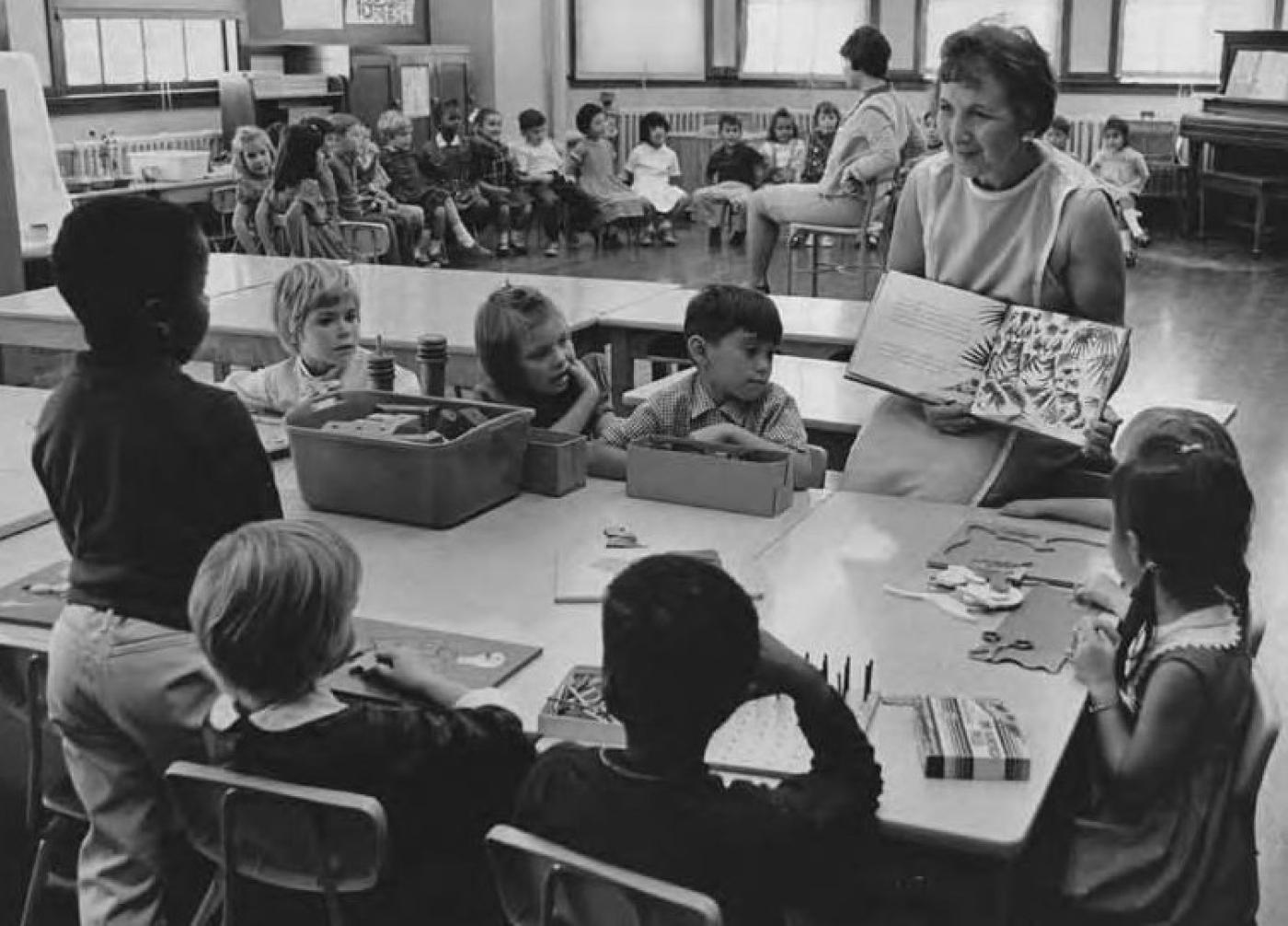 Sara Efron reading and showing book to children seated around table in classroom at Bailey Gatzert School with other group of children and teacher in background, 1966